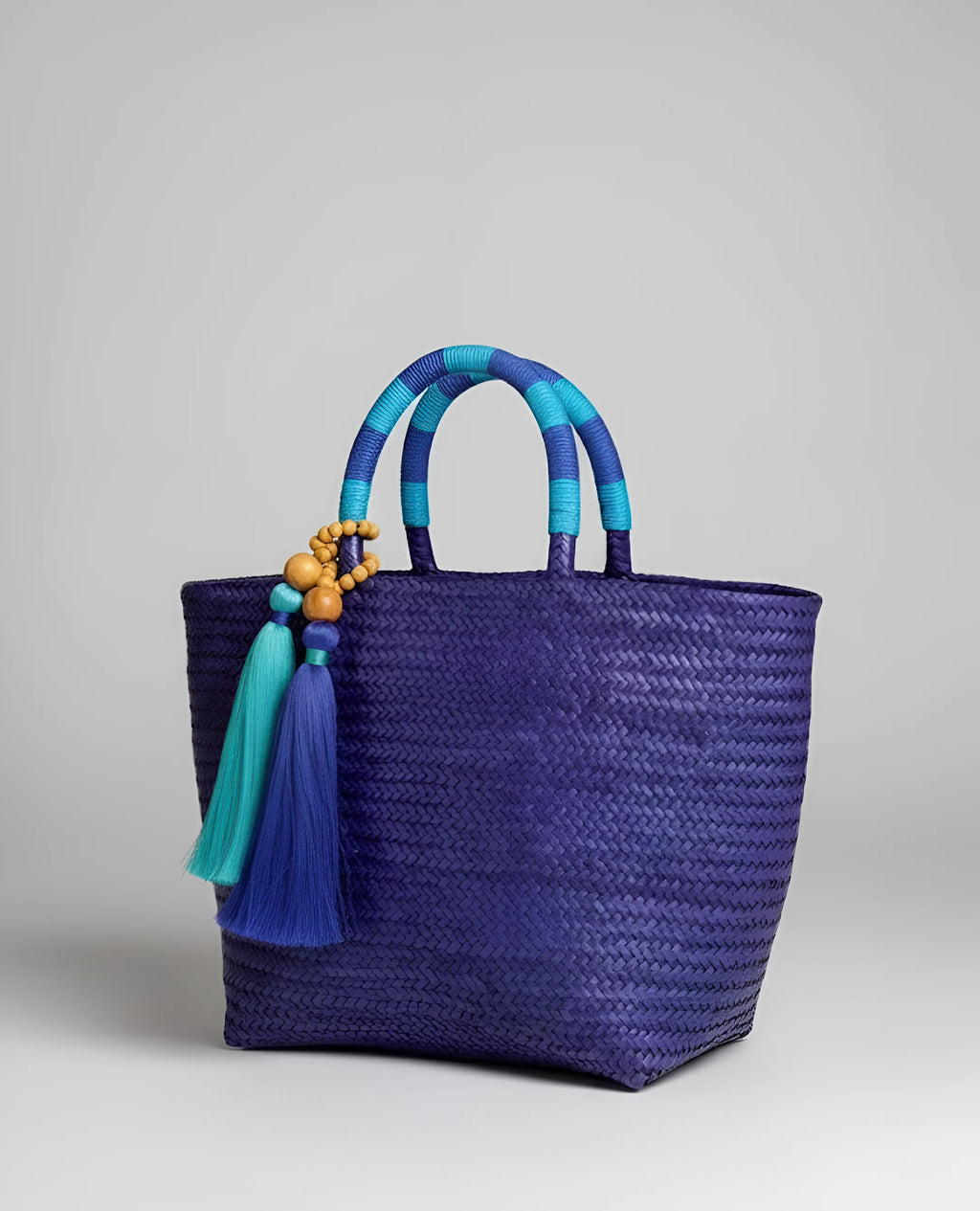 Brazilian handcrafted straw bag with dark blue and purple woven texture, tassel details in aqua green, and elegant coastal resort style.