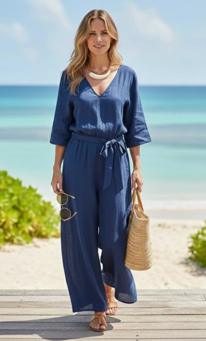 Woman in a blue jumpsuit standing on a beach with ocean and sky in the background wearing a horn necklace.